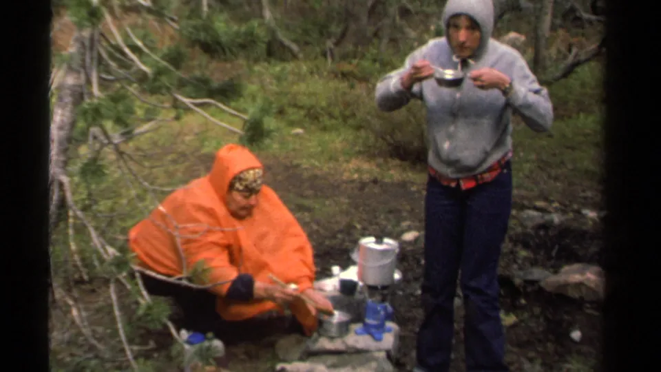 A person in an orange poncho crouches by cookware while another in a gray hoodie drinks from a metal bowl in the Lake Tahoe woods.