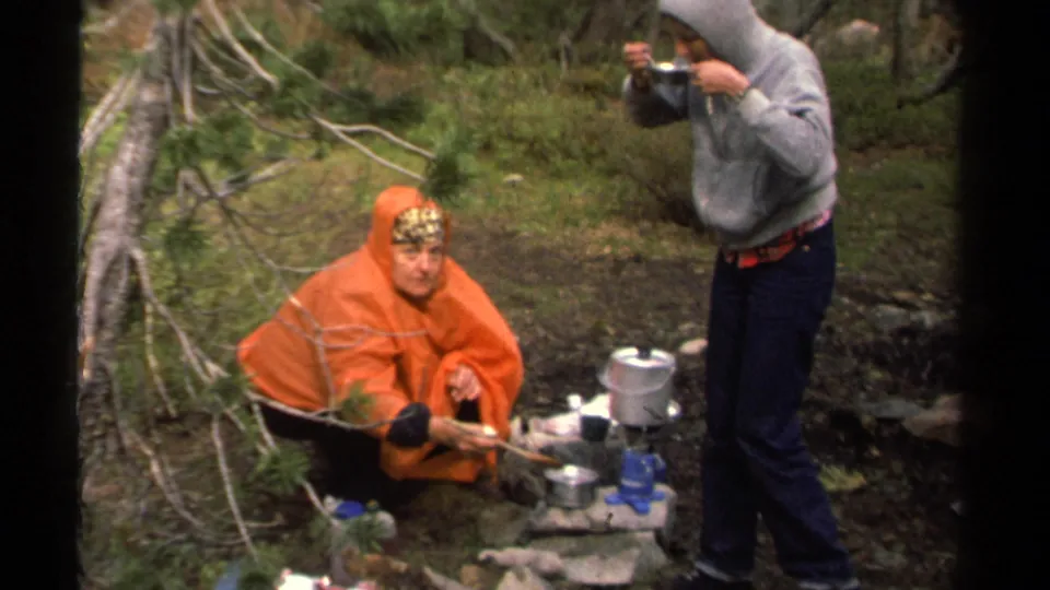The 1978 8mm frame shows a crouching camper in an orange poncho tending a pot while a standing companion eats or drinks nearby.