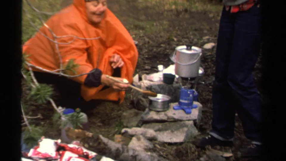 A closer Lake Tahoe home-movie frame shows a person in an orange poncho handling camp cookware beside a blue stove and silver pot.