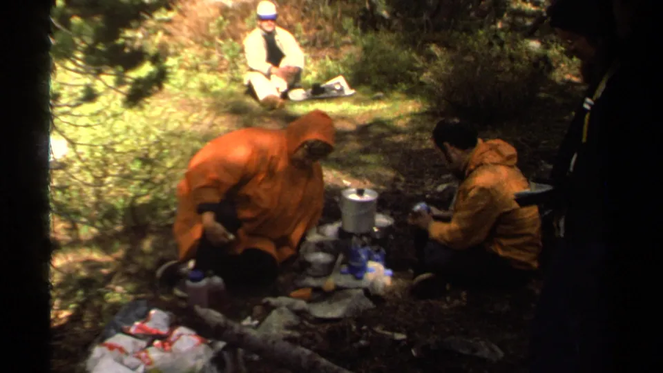 Campers near Lake Tahoe appear to prepare a meal around a compact stove, with pine cover and dappled sunlight around them.