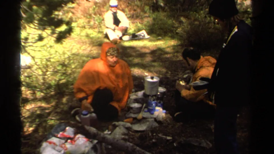 Home-movie frame of a 1978 Lake Tahoe campsite with an orange-ponchoed camper, stove, supplies, and companions nearby.