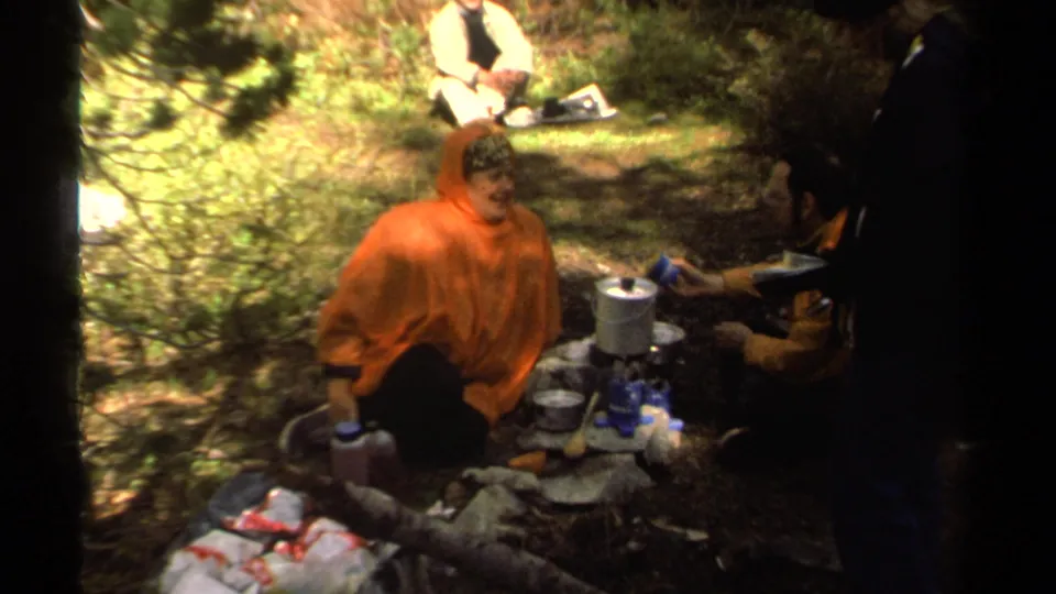 A camper in an orange poncho looks toward the camera as companions continue preparing food at a wooded Lake Tahoe campsite.
