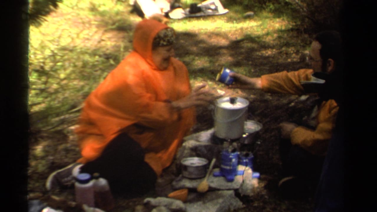 Camper in an orange poncho sits beside a small stove and metal pot in the woods.