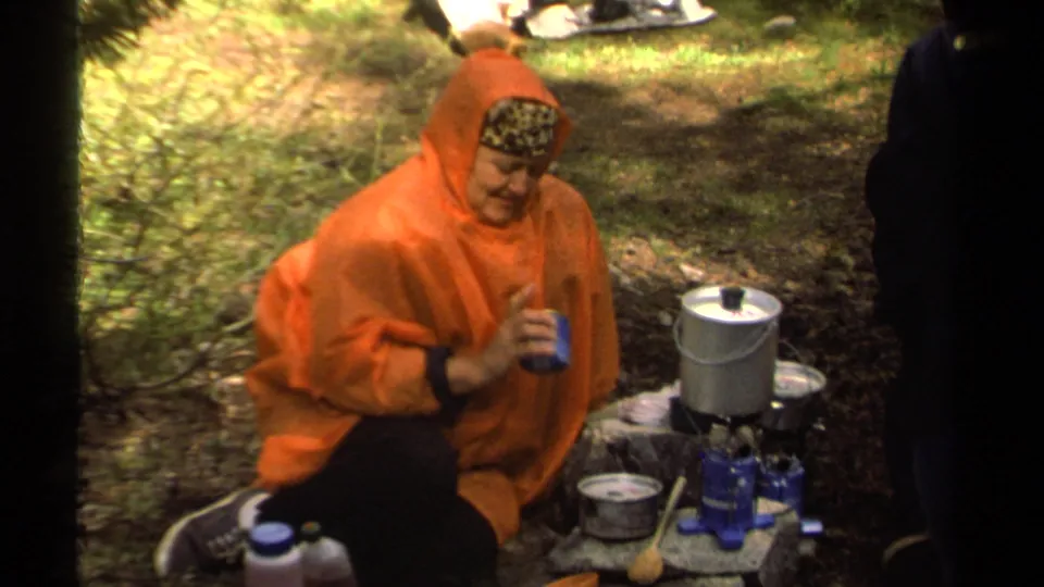 A camper in an orange poncho drinks from a blue cup near a metal pot and small stove in the wooded Lake Tahoe camping scene.