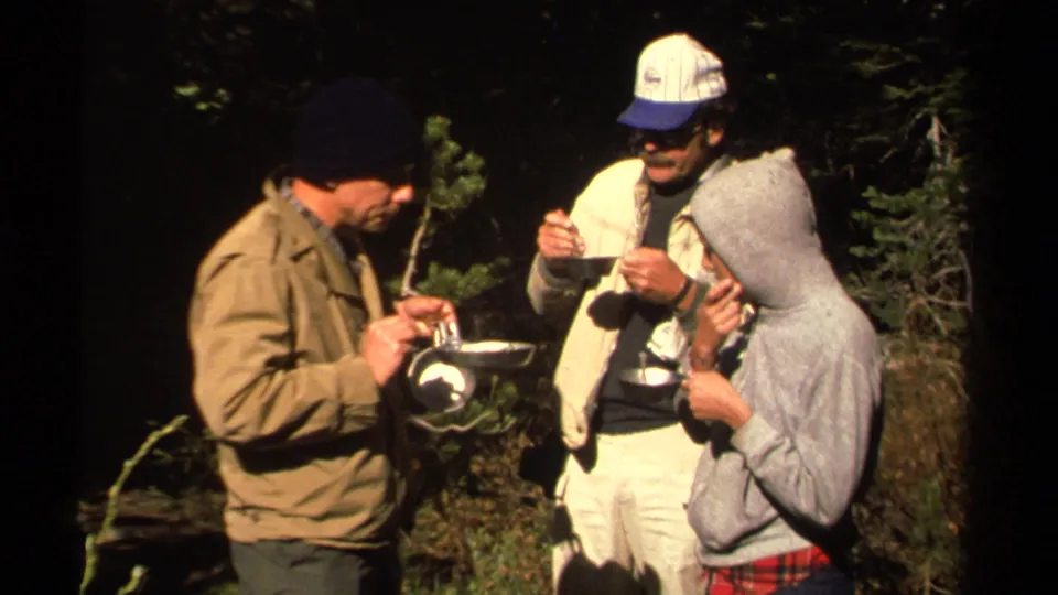 Three adults in jackets, caps, and a hooded sweatshirt eat from bowls and cups in 1978 Lake Tahoe-area camping footage.