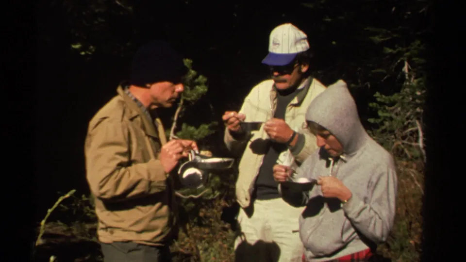 A small group of campers in layered clothing eat from bowls together in the wooded Lake Tahoe-area campsite scene.