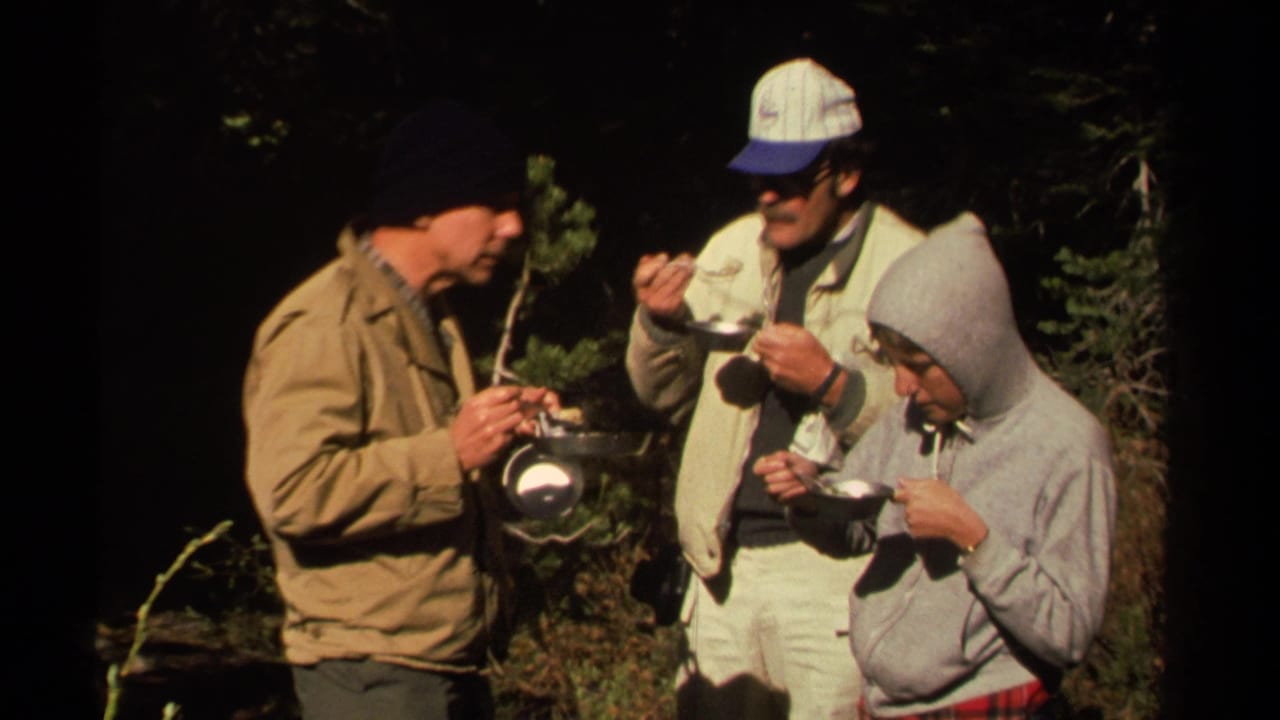 Three men stand in woods eating from bowls and cookware.