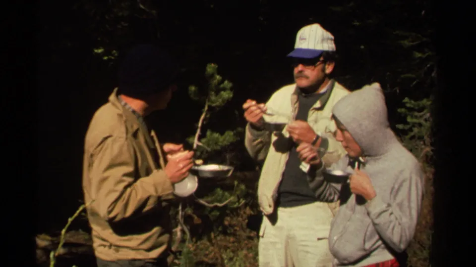 Three adult men in casual outdoor clothing eat from camp bowls in a shaded Lake Tahoe forest setting.