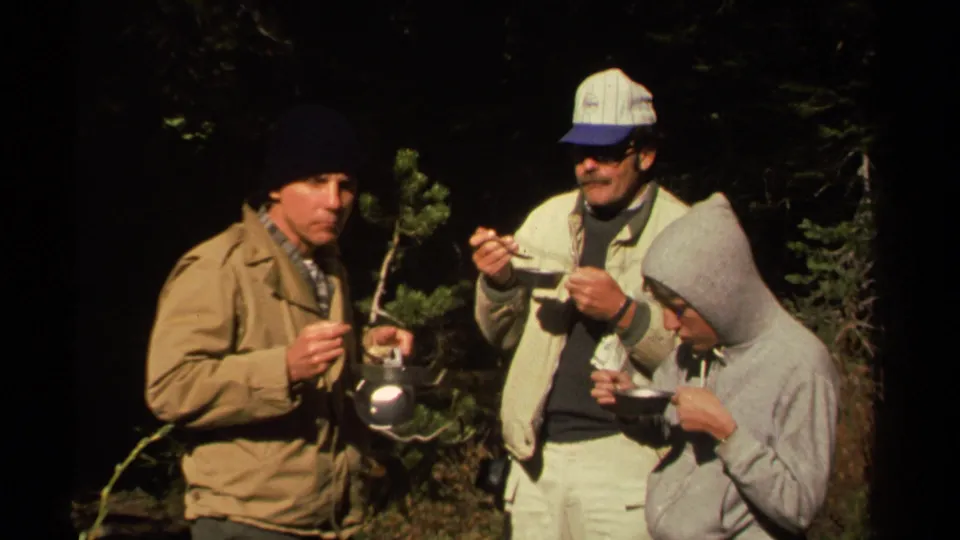 A man in a tan jacket holds a metal pot while two companions eat during a 1978 wooded camping meal.