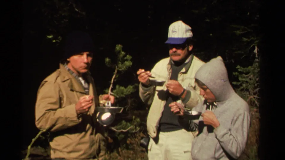 Three adult men continue a simple campsite meal in the woods, with bowls, spoons, and aluminum cookware visible.