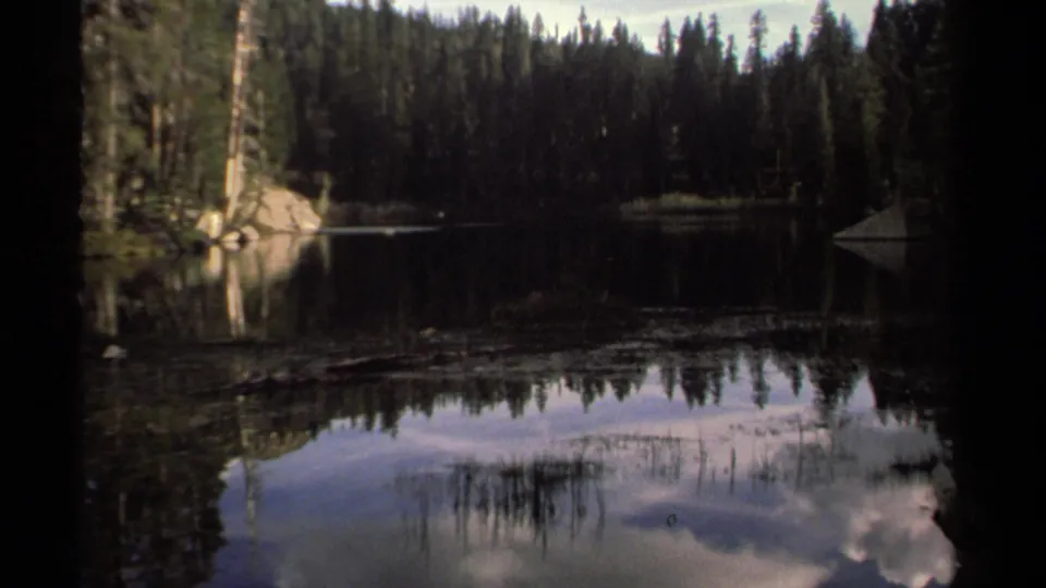 1978 Lake Tahoe home-movie frame showing pine trees, shoreline rocks, and cloud reflections on still water.