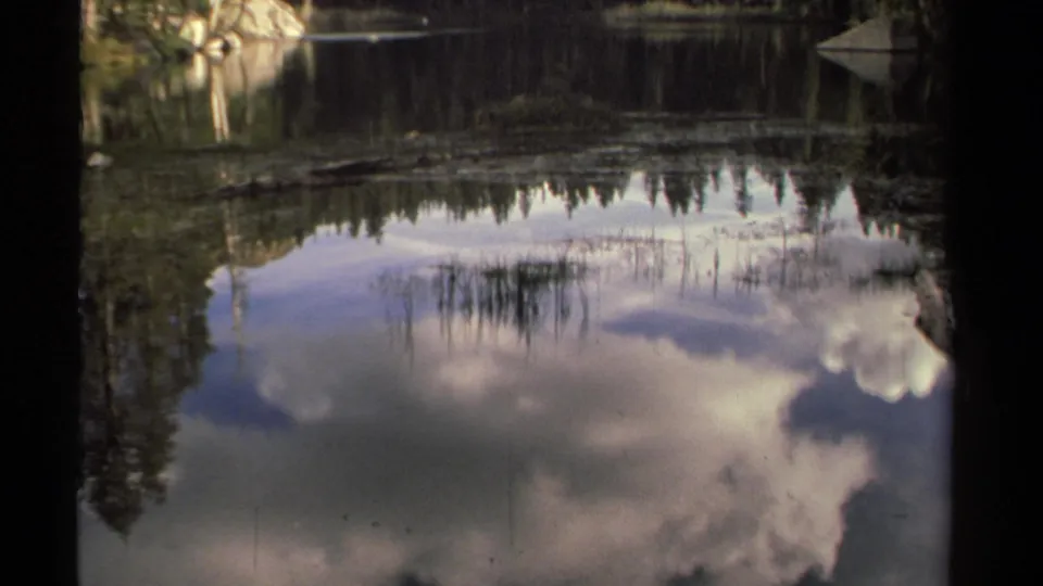 8mm frame of a Lake Tahoe creek or lake edge, with reflected clouds, trees, and shoreline rocks in soft vintage color.