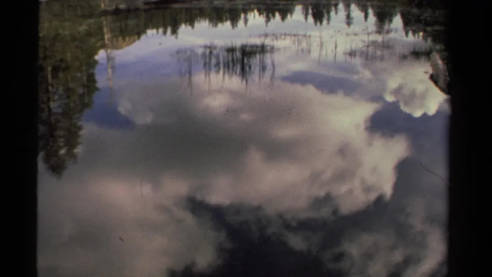 Late-1970s Lake Tahoe home-movie still emphasizing cloud reflections, dark water, reeds, and a pine-lined shore.