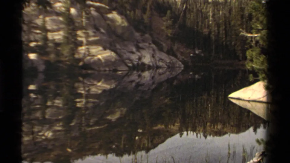 1978 Lake Tahoe landscape frame showing still water reflecting rocky mountain slopes and pines, with a pale shoreline boulder at right.