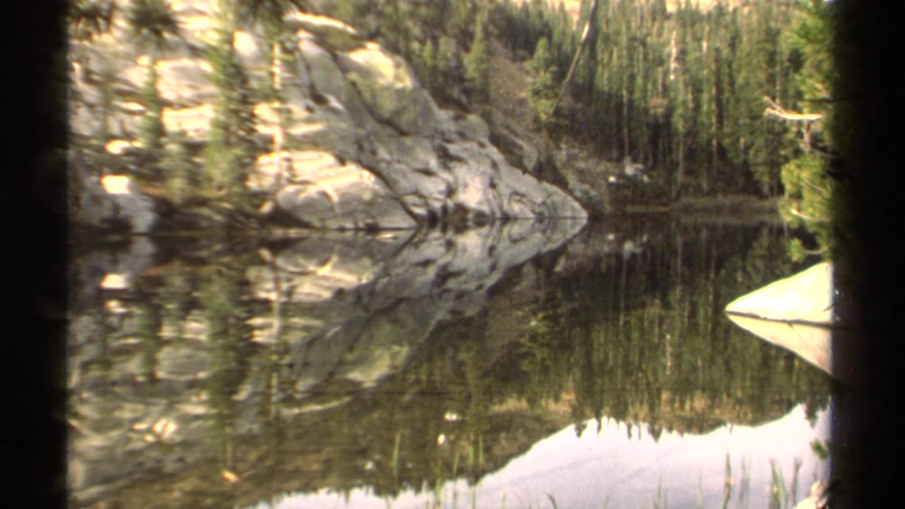 1978 Lake Tahoe 8mm frame showing calm water mirroring a rocky forested shoreline and pale sky.