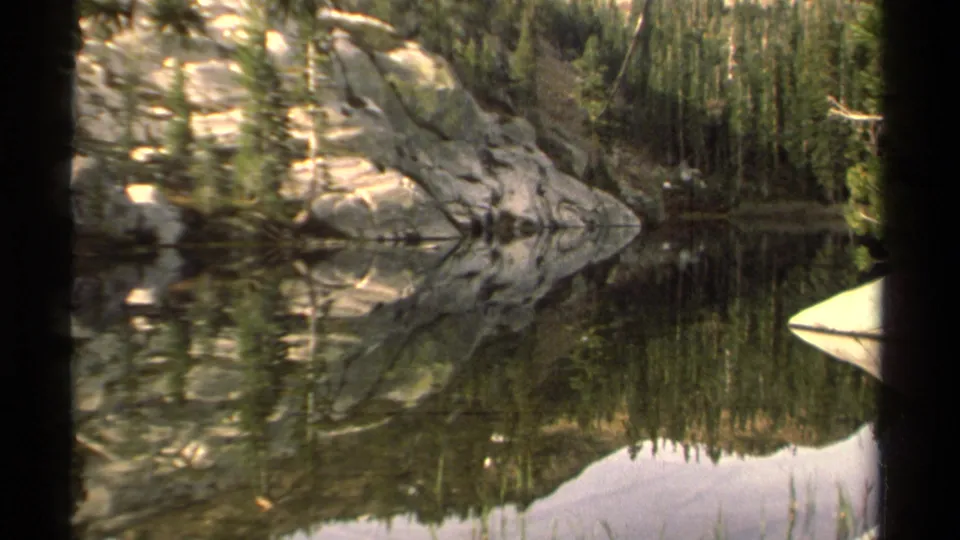 Lake Tahoe shoreline on 8mm film, with pale rocks and conifers reflected in glassy water.