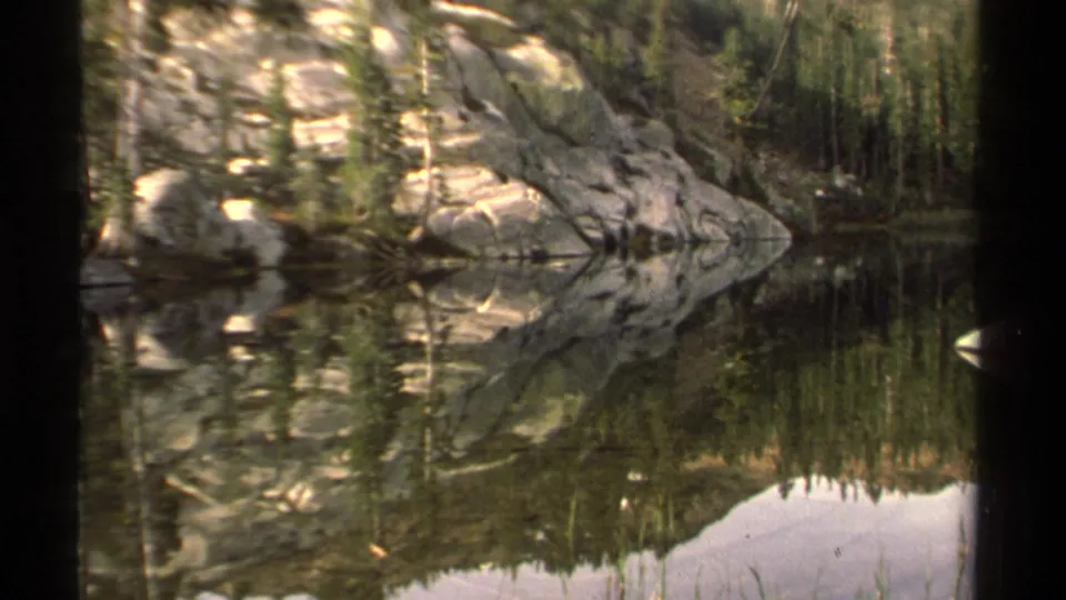 1978 home-movie view of a quiet Lake Tahoe inlet with trees and granite reflected in the lake.