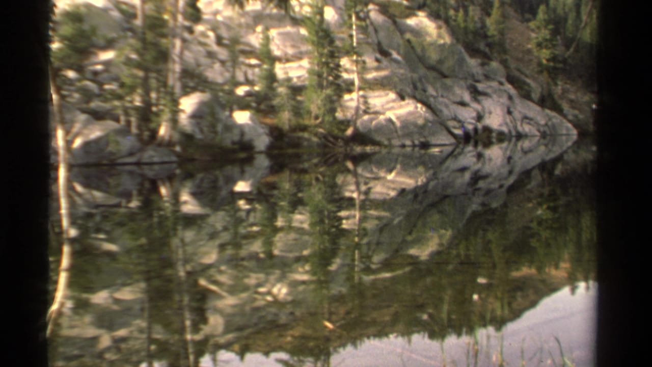 Calm lake water reflecting evergreen trees and pale rocks along a rocky shoreline.