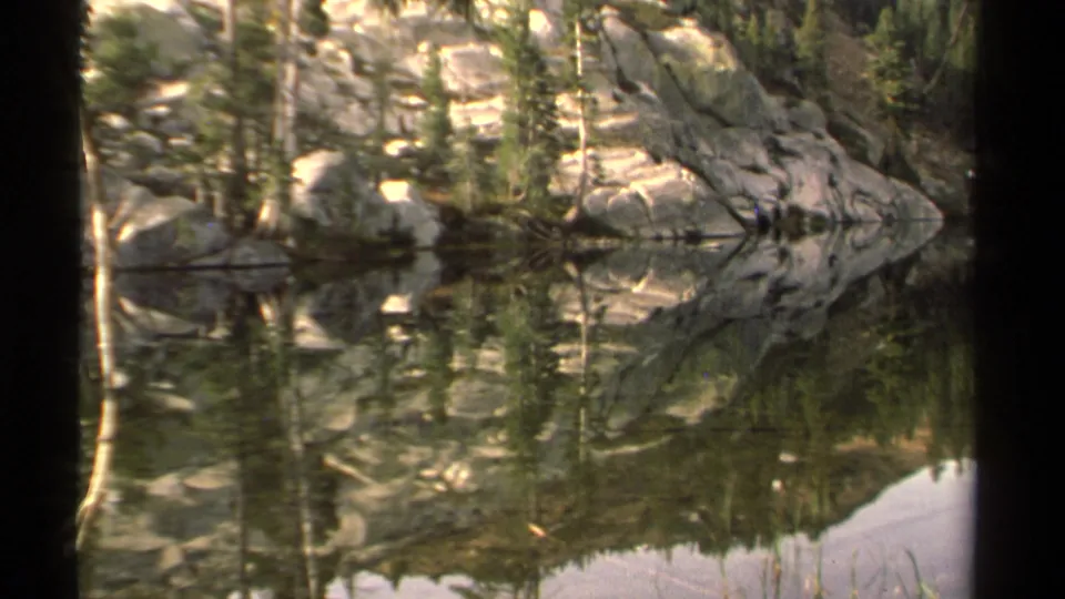 1978 Lake Tahoe home-movie frame showing still water with mirrored evergreen trees, rocks, and shoreline in soft 8mm grain.