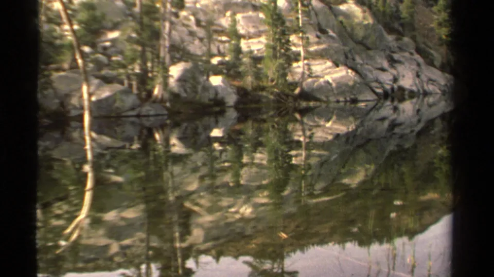Lake Tahoe 8mm frame with rocky shore, evergreen trees, and their reflections across calm water, partly bordered by a dark film edge.