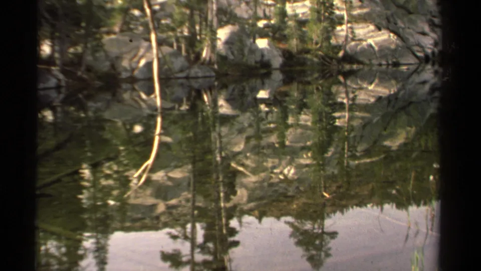 Lake Tahoe home-movie frame with evergreen and rock reflections, a pale sky patch on the water, and soft 8mm texture.