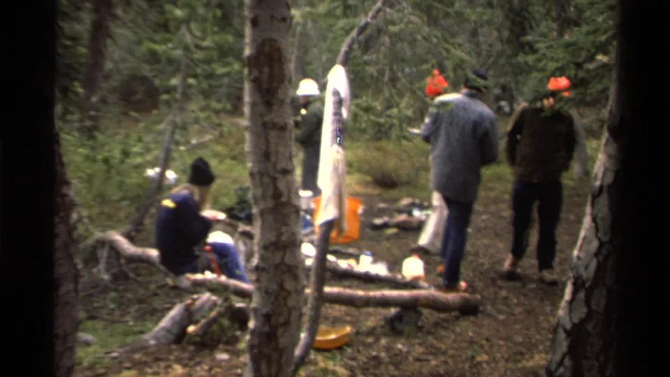 1978 Lake Tahoe home-movie frame showing campers among pine trees, with one person seated on a fallen log eating while others stand nearby.