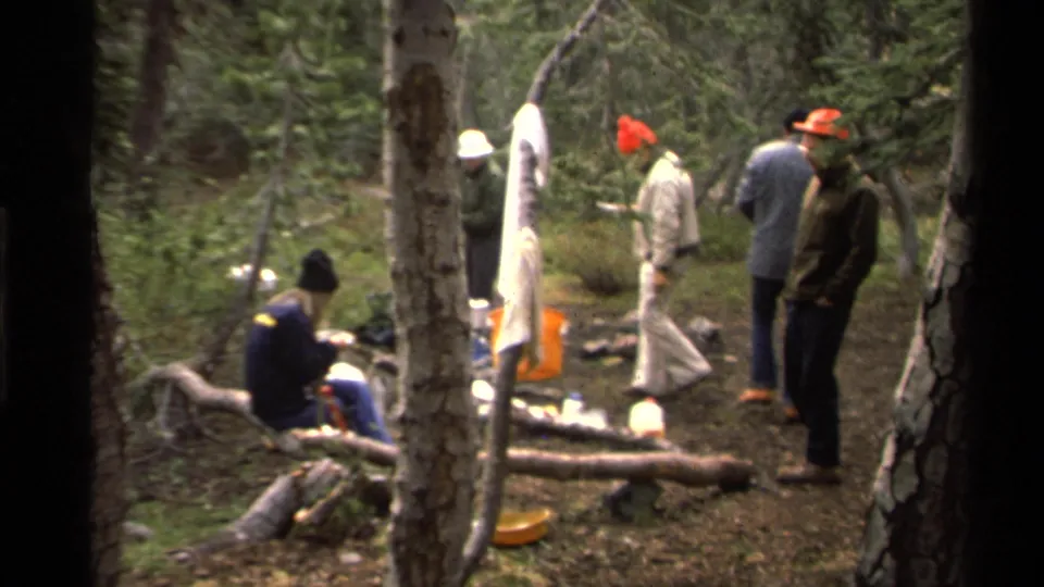 1978 Lake Tahoe camping scene with a seated person eating on a log and several people in jackets and bright hats standing nearby.