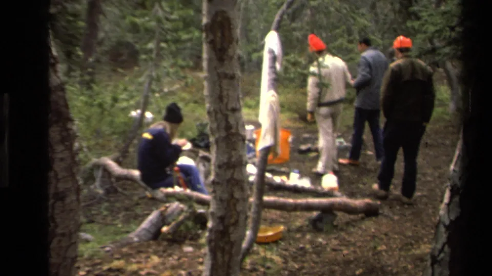 1978 Lake Tahoe home-movie frame of campers standing and eating around a rough forest campsite framed by dark tree trunks.