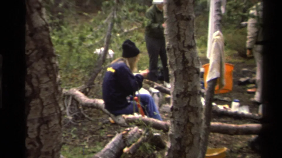 1978 Lake Tahoe frame showing a camper in a dark jacket and knit cap eating from a plate on a log, with gear and other campers nearby.