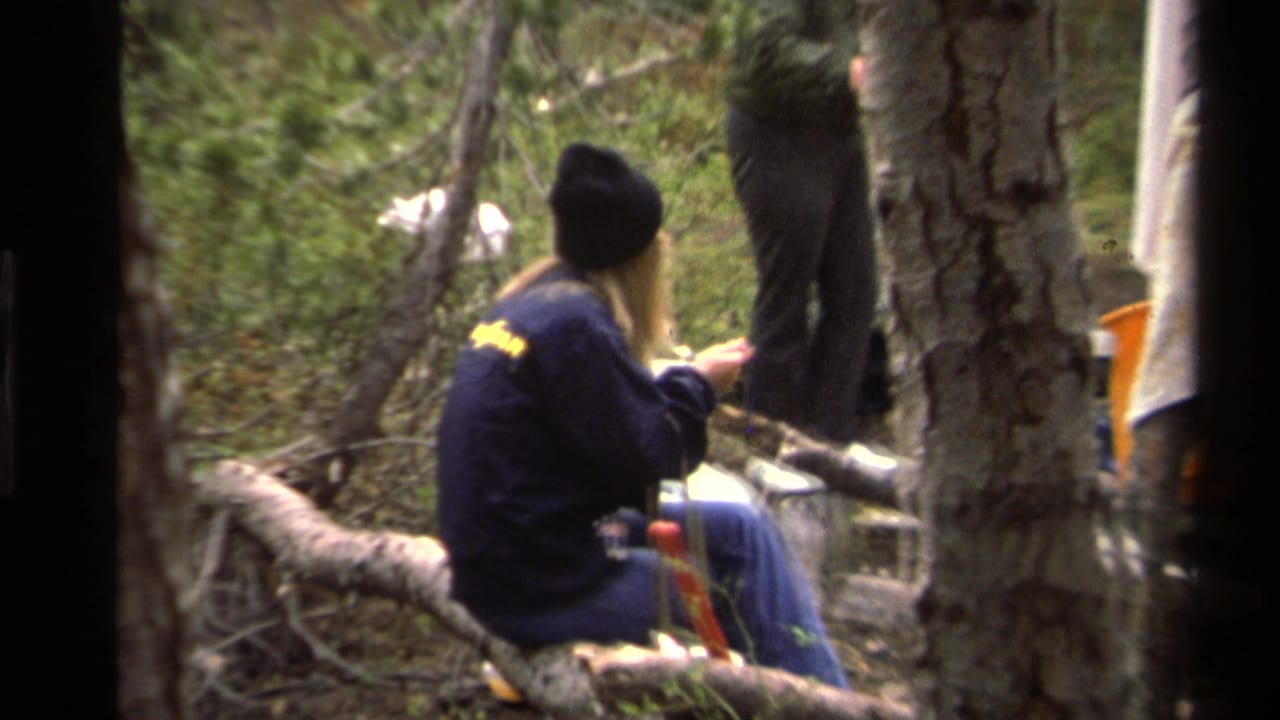 Young woman in a dark jacket and beanie sits on a fallen log in a wooded campsite.