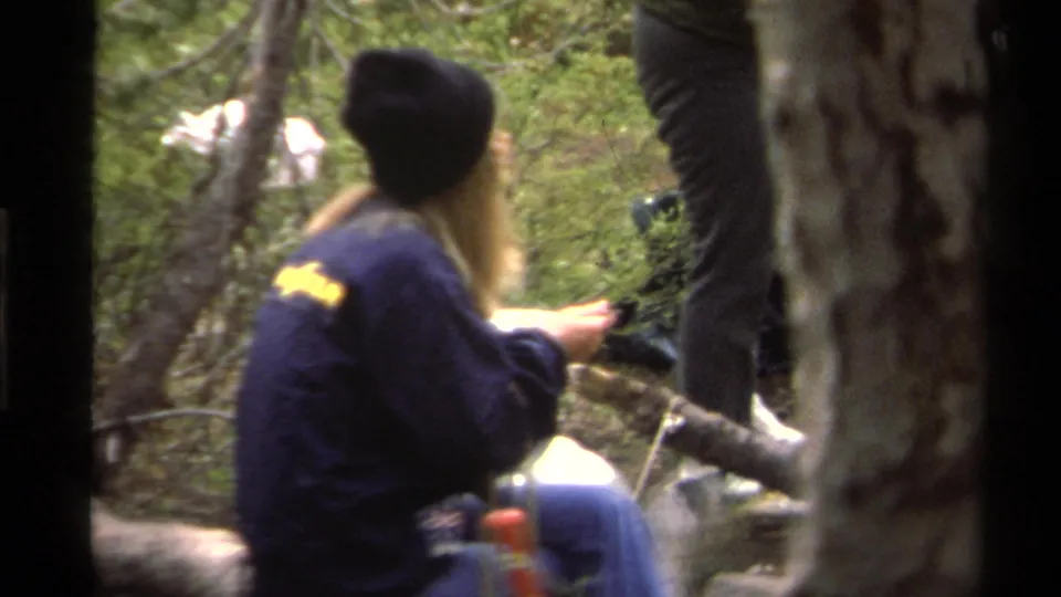 A camper in a dark jacket and beanie sits on a fallen log in a wooded Lake Tahoe campsite, with other adults partly visible nearby.