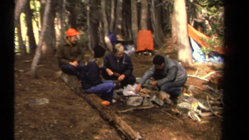 A group of adult campers sits in a wooded Lake Tahoe campsite near supplies and a bright tent, filmed in soft 8mm color.