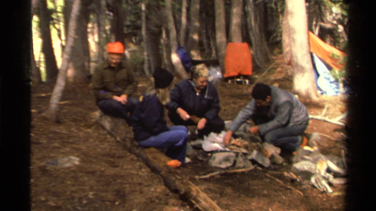 Four adults at a 1978 Lake Tahoe forest campsite sit around a small fire area, with camping bags, logs, trees, and an orange shelter visible.