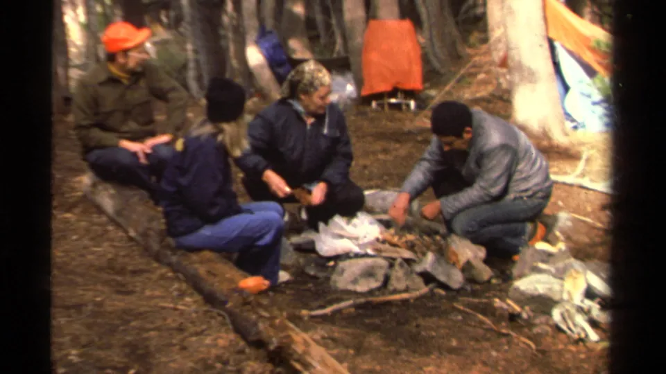 Four adults sit at a forest campsite near Lake Tahoe, with a gray-jacketed camper handling materials by the small fire area.