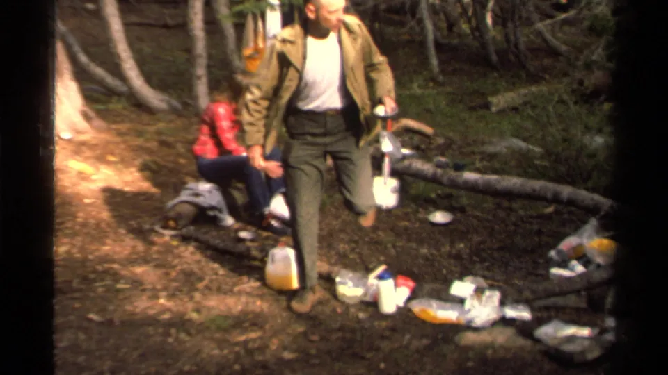In the Lake Tahoe campsite scene, a man carries a shovel past scattered camp supplies while a woman in red sits on a log.