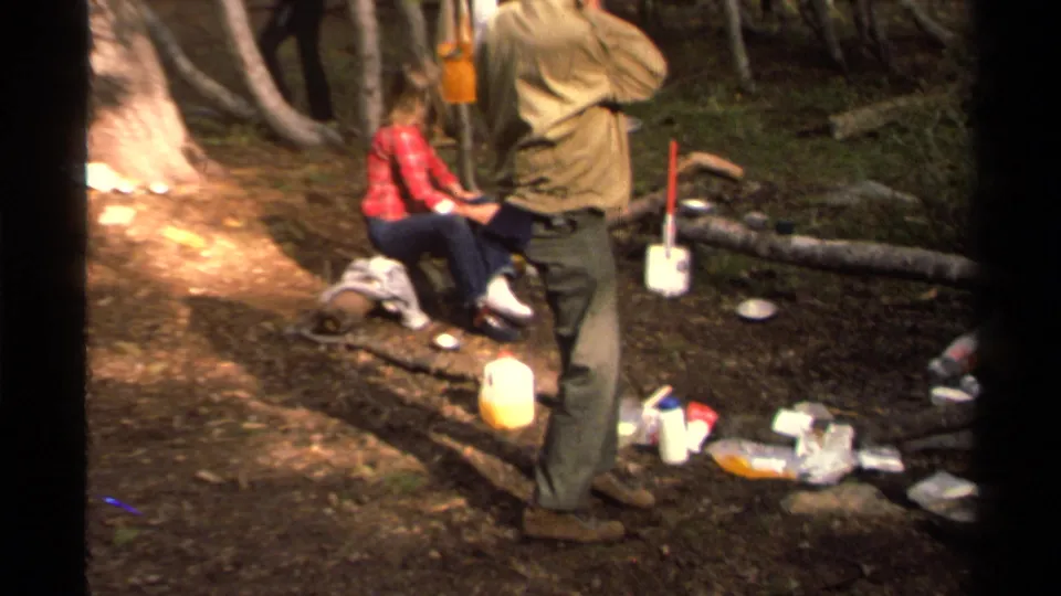 The handheld 8mm frame shows a man standing near a yellow jug and shovel while a woman sits on a log in the forest camp.