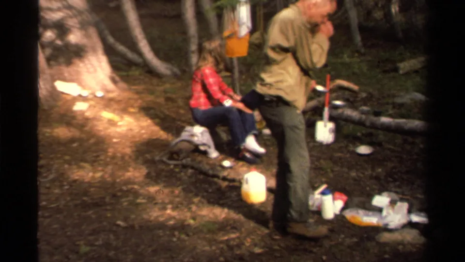 At the forest campsite, a man in a tan jacket pauses near a shovel and yellow jug as a woman sits on a log nearby.
