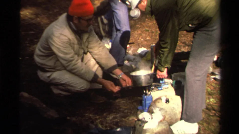 A man in a red knit cap and another hiker work beside a small stove and pot in 1978 Lake Tahoe footage.