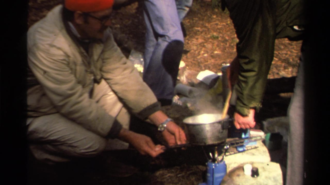 Adults crouch around a blue camp stove and metal pot on a forest floor.
