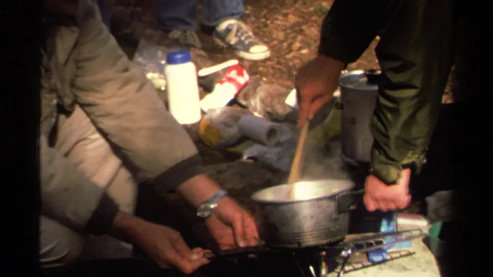 1978 Lake Tahoe camping footage frame with a wooden spoon stirring a steaming metal pot over a compact blue stove.