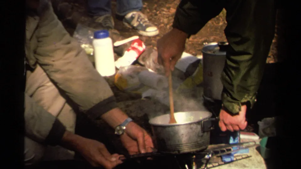 Close Super 8 frame from Lake Tahoe showing hands near a stove, a steaming pot, and camping supplies during outdoor cooking.
