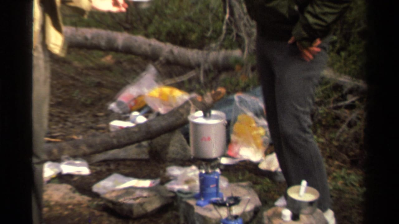 1978 Lake Tahoe campsite frame showing a small blue stove, silver pot, scattered bags, and the lower body of an adult camper near a fallen log.