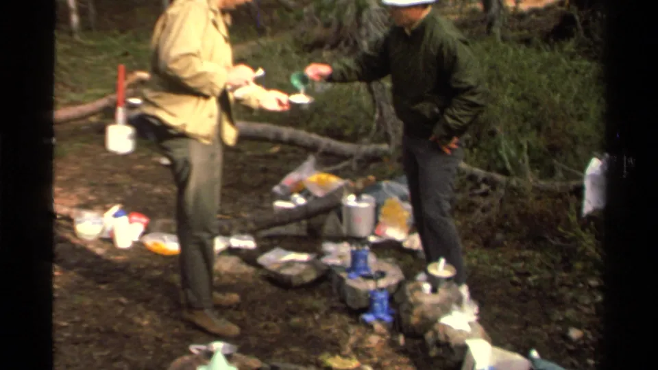 Two adults at a 1978 Lake Tahoe wooded campsite handle a cup and bowl near scattered meal supplies and a compact camp stove.