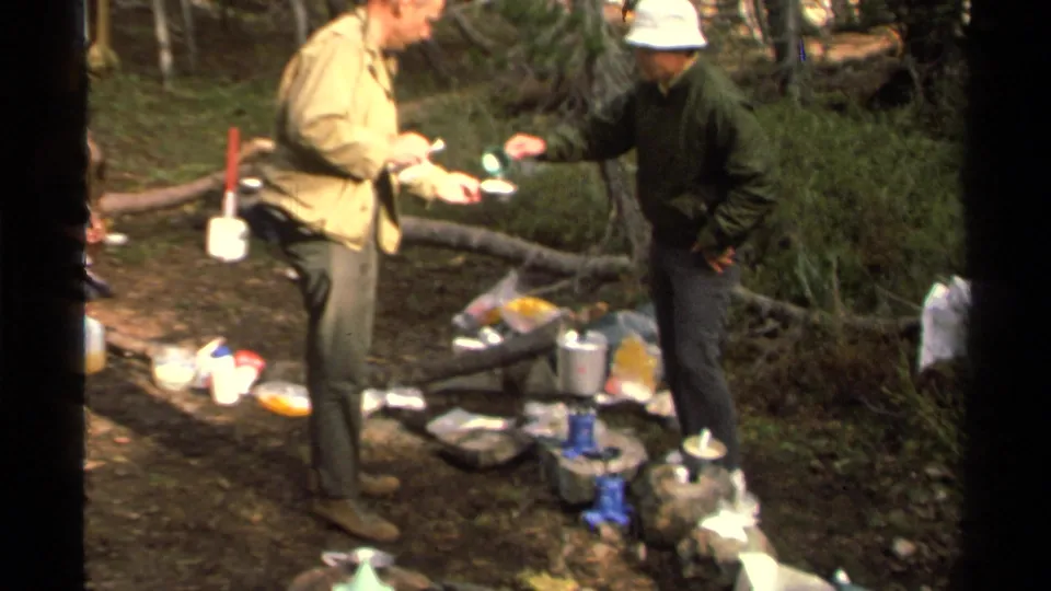 1978 Lake Tahoe home-movie frame of two adult campers preparing or sharing food and water among pots, bags, rocks, and forest undergrowth.