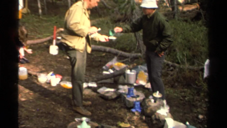 Two adults in 1978 Lake Tahoe camping footage stand near a blue stove and silver pot while handling a cup and bowl on the forest floor.