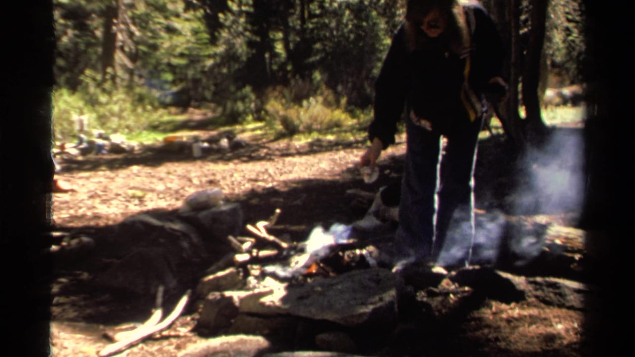 Woman standing near a small smoky campfire in a wooded area.