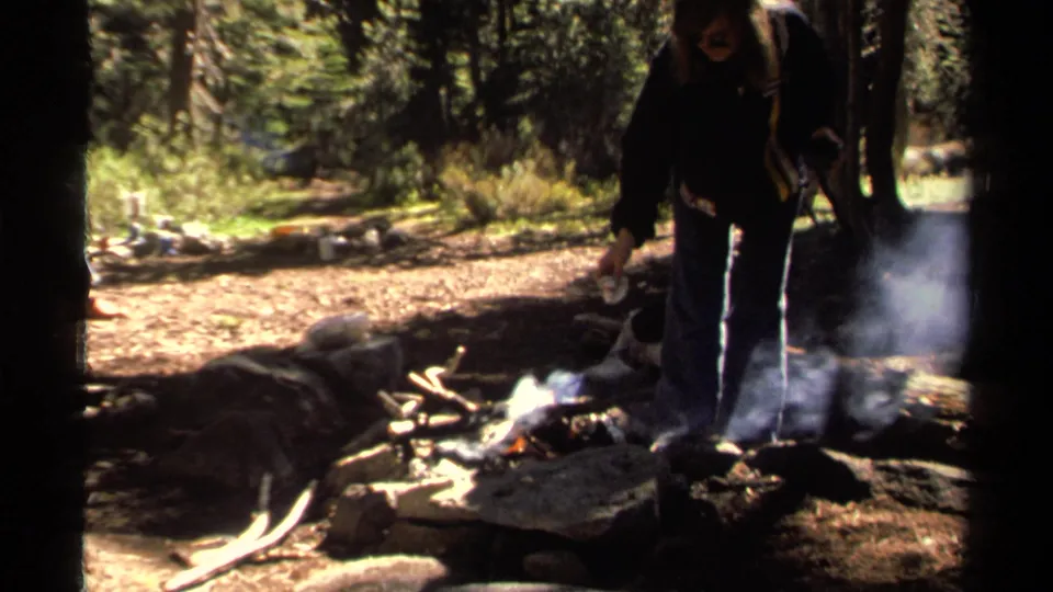 A woman stands beside a smoking campfire in a wooded Lake Tahoe setting, with rocks and kindling nearby.