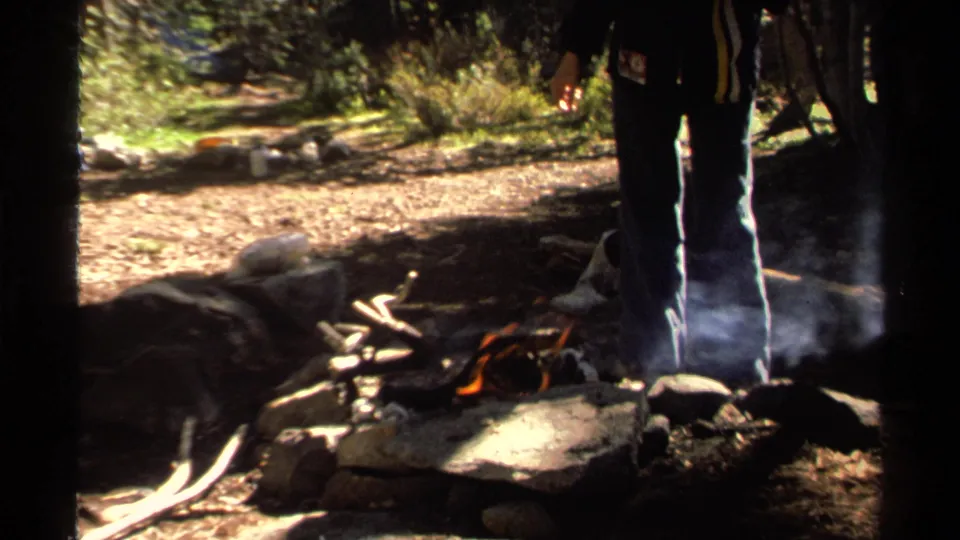 The frame shows the lower body of a woman near a smoking campfire, with kindling and rocks on the forest floor.