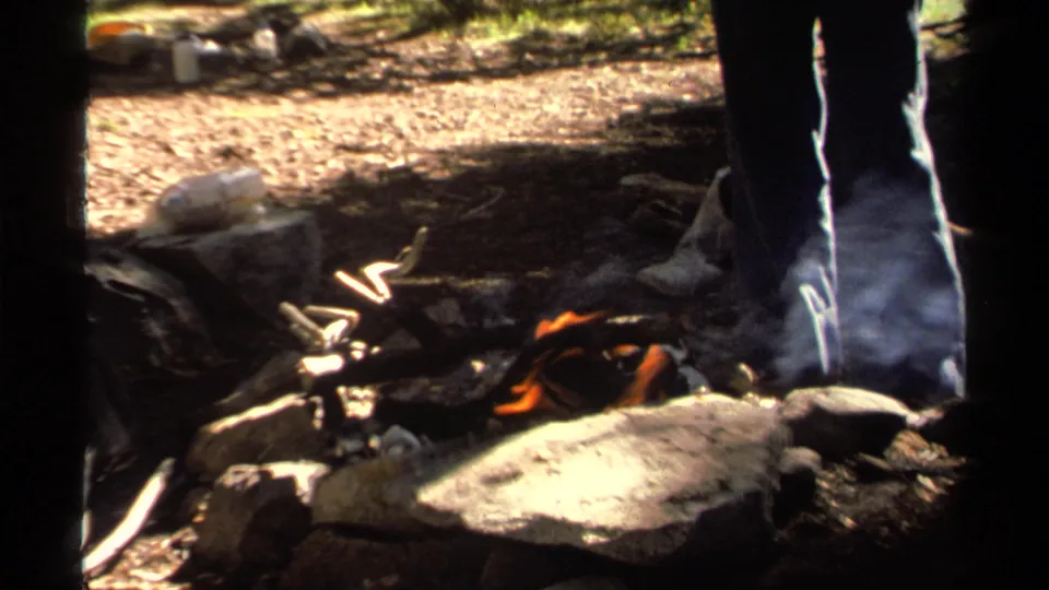 A small campfire burns and smokes beside rocks while a woman stands nearby in the Lake Tahoe woods.