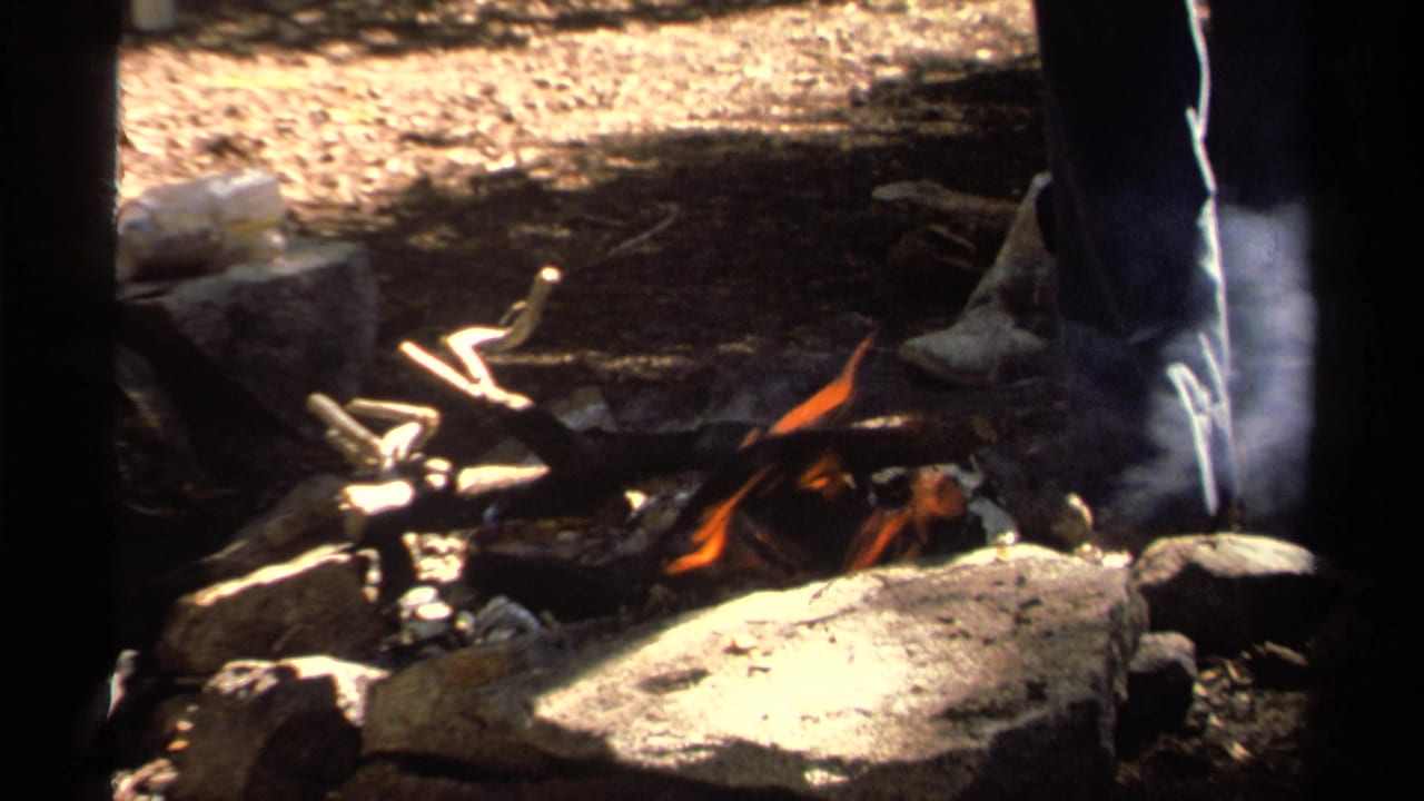 1978 Lake Tahoe 8mm frame showing a small campfire among stones, logs, smoke, and the lower legs of a nearby person.
